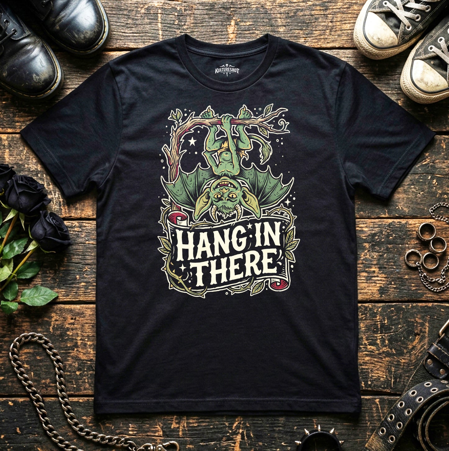 Black t-shirt with 'Hang'in There' graphic design on a wooden surface with shoes and chains.