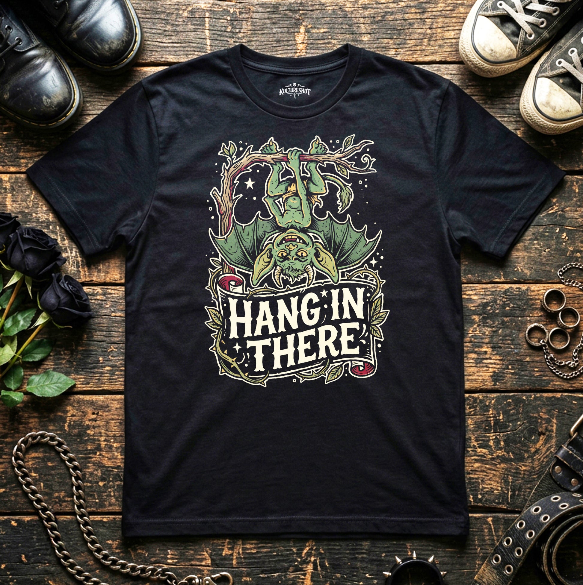 Black t-shirt with 'Hang'in There' graphic design on a wooden surface with shoes and chains.