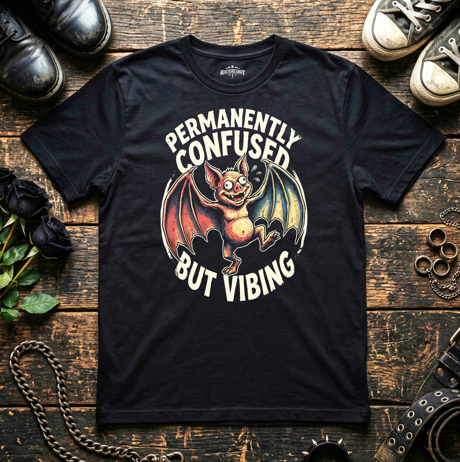 Black t-shirt with a bat graphic and text 'Permanently Confused But Vibing' on a wooden surface with shoes and chains.