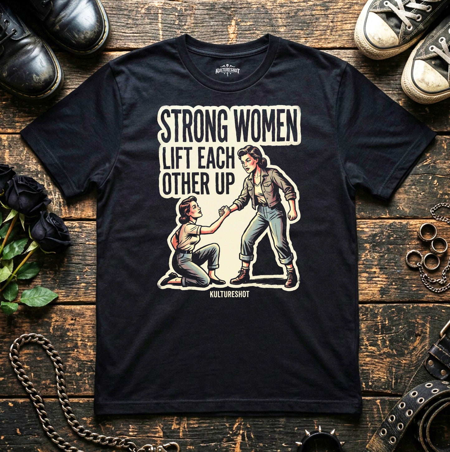 Black t-shirt with graphic design and text 'Strong women lift each other up' on a wooden surface with shoes and plants.