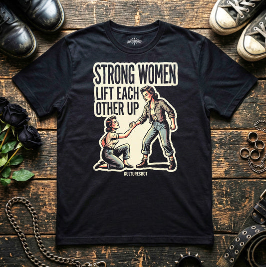 Black t-shirt with graphic design and text 'Strong women lift each other up' on a wooden surface with shoes and plants.