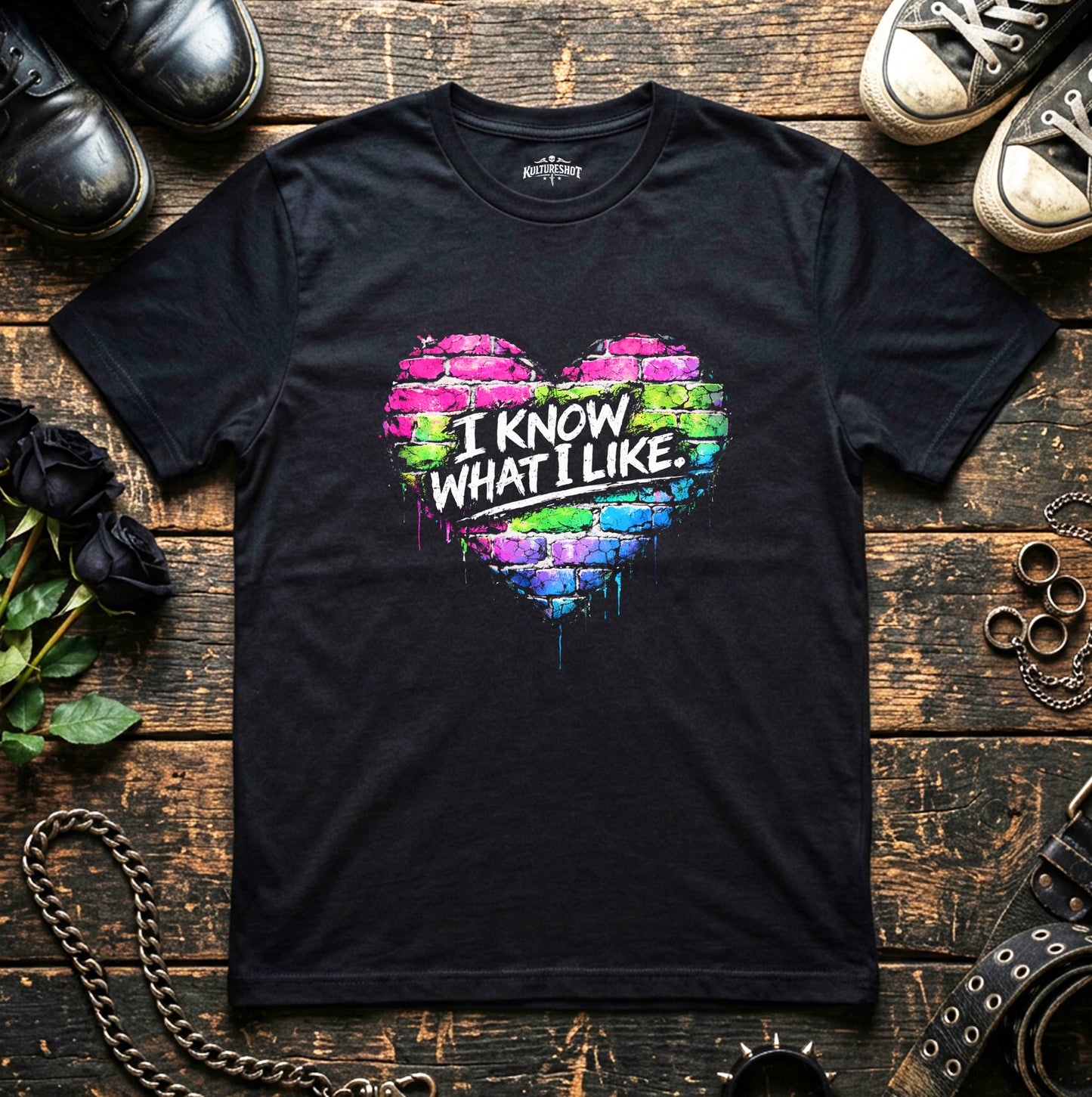 Black t-shirt with colorful heart design and text on a wooden surface with shoes and jewelry.