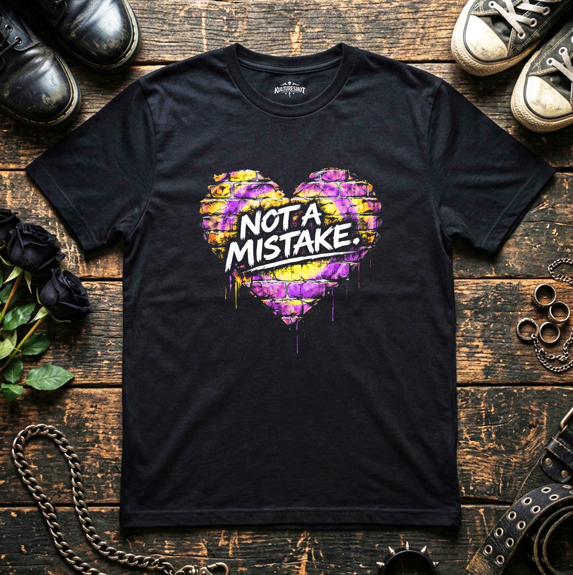 Black t-shirt with colorful heart design and 'Not a Mistake' text on a wooden surface with shoes and flowers.