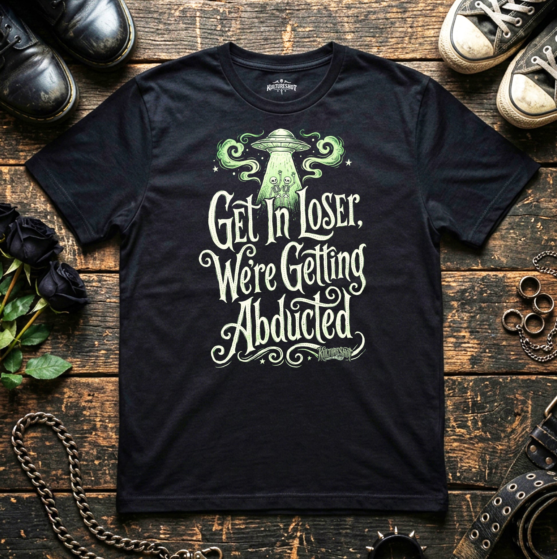 Black t-shirt with alien abduction graphic and text on a wooden surface with shoes and plants.