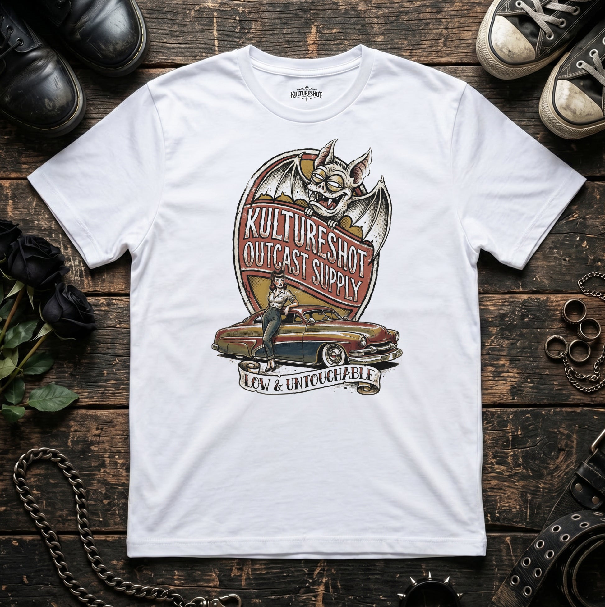 White t-shirt with a graphic design on a wooden surface with shoes and accessories.