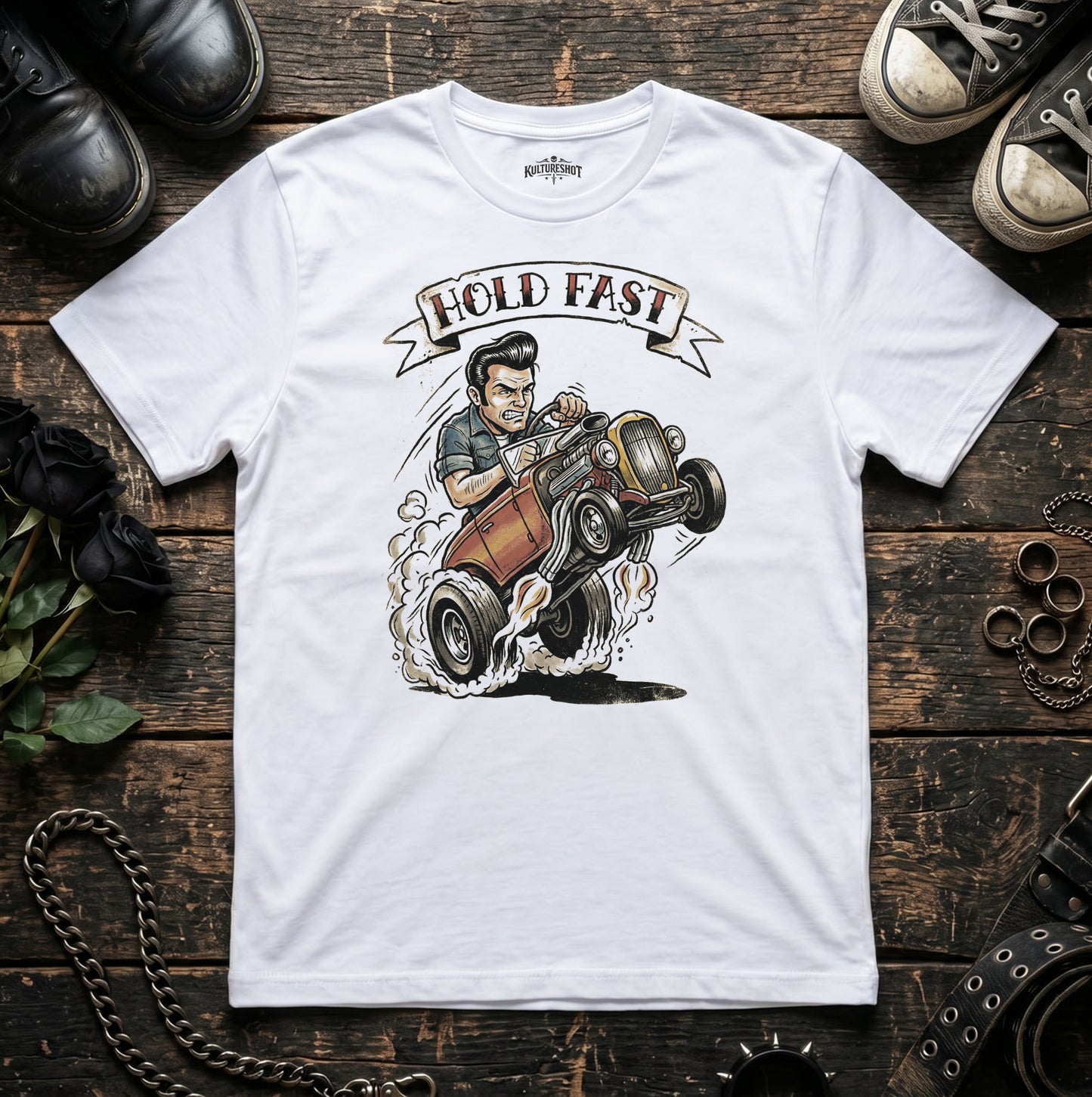 White t-shirt with a graphic of a man driving a hot rod and 'Hold Fast' text, placed on a wooden surface.