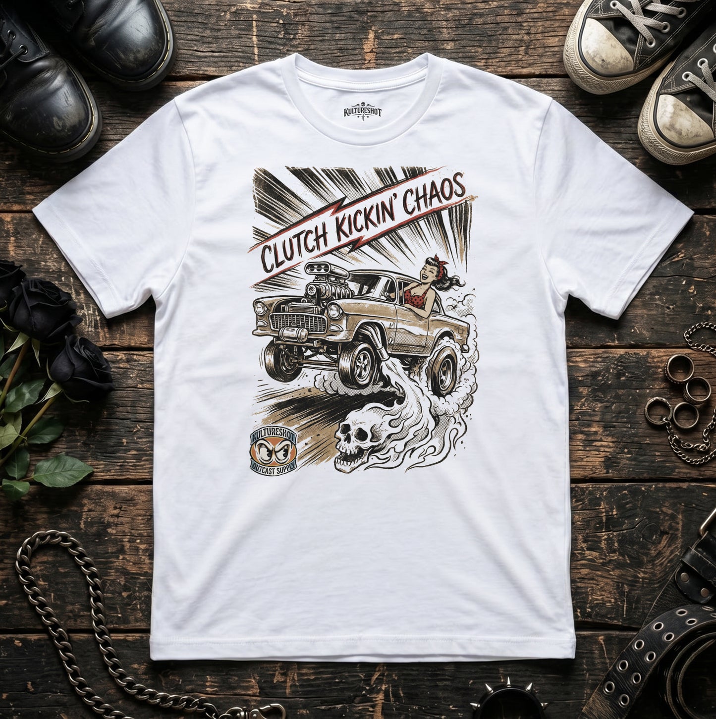 White t-shirt with a graphic design of a car and text 'Clutch Kickin' Chaos' on a wooden surface.