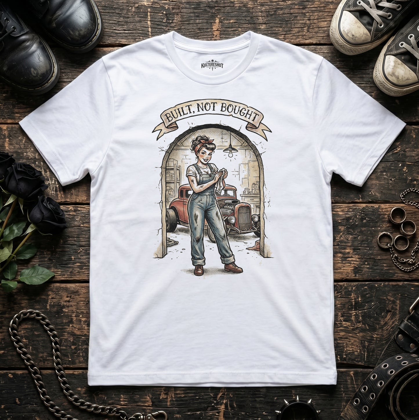 White t-shirt with graphic design of a woman in overalls and text 'Built, Not Bought' on a wooden surface with shoes and tools.
