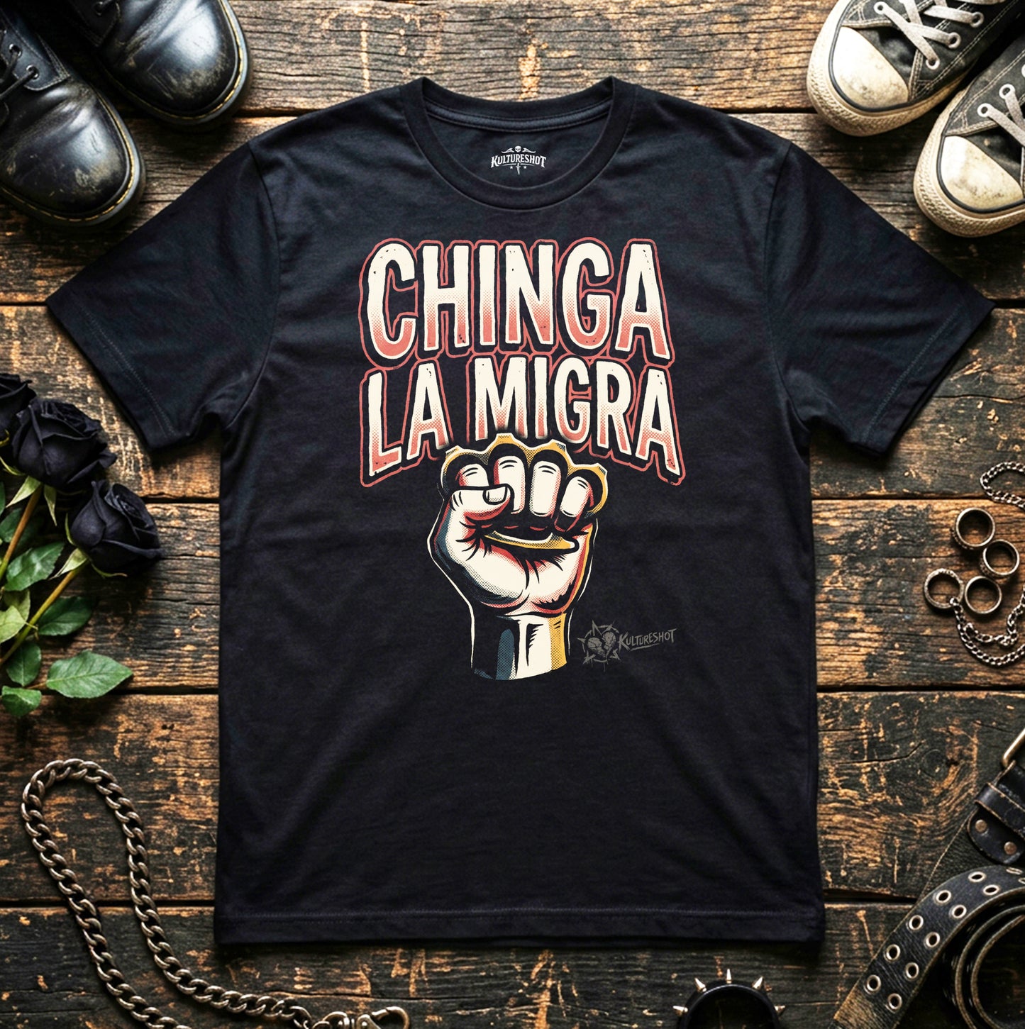 Black t-shirt with 'Chinga La Migra' graphic on a wooden surface with shoes and plants.