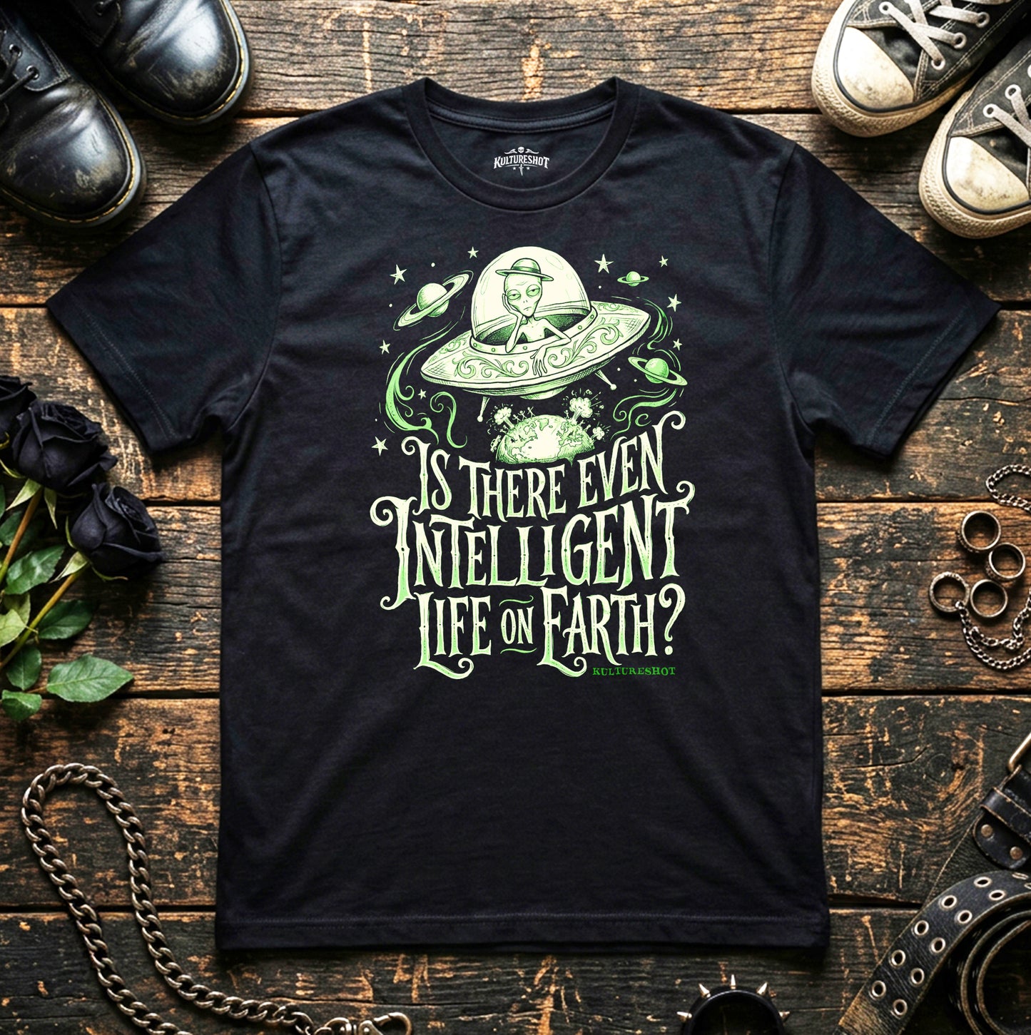 Black t-shirt with alien abduction graphic and text on a wooden surface with shoes and plants.